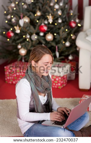 Pretty young woman, laughing while chatting to online friends using laptop computer, near Christmas tree decoration with lights.