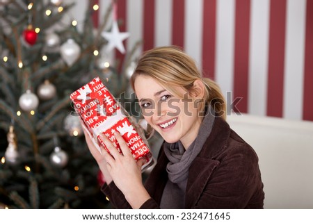 Young woman trying to guess the contents of a Christmas gift holding it to her ear as she shakes it and smiles at the camera