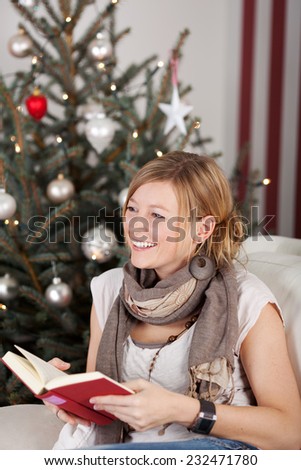 Happy woman relaxing at Christmas with a book as she sits on a sofa in front of the tree laughing happily at someone off frame