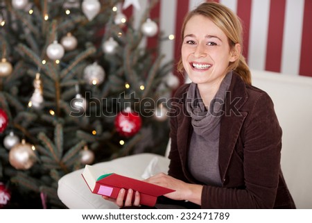 Pretty woman relaxing in front of a Christmas tree reading a book and pausing to look at the camera with a lovely smile