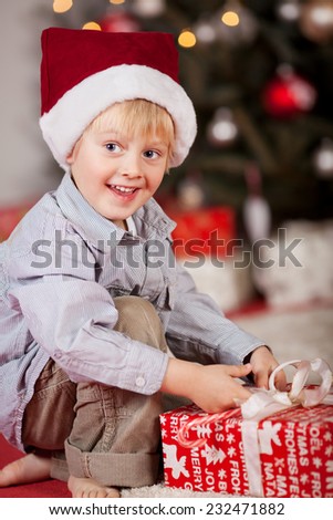 Happy smiling little boy unwrapping a Xmas gift as he kneels bare foot on the floor in front of the Christmas tree in his festive red Santa hat