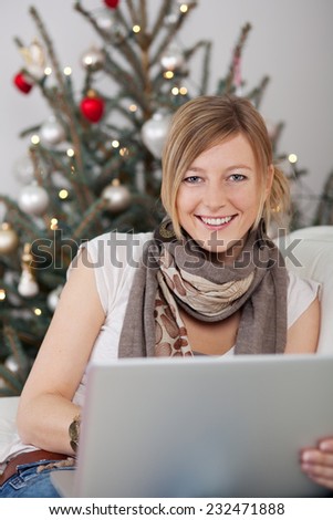 Pretty Smiling Woman with Brown Neck Scarf Sitting on White Couch Holding Laptop While looking at the Camera with Christmas Tree Decoration at the Background.
