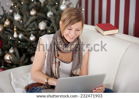 Happy Pretty Woman in Casual Clothing Sitting on White Couch While Browsing Something Using her Laptop Computer. Captured Indoor with decorated Christmas Tree at the Back.