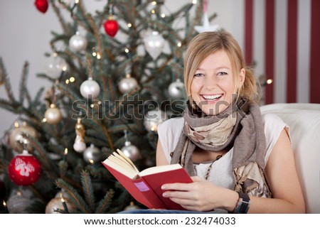 Happy young woman reading a book at Christmas as she sits in front of the Christmas tree looking at the camera with a lovely warm smile