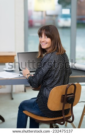 Young woman working at a table in the office at a laptop computer turning in her chair to smile at the camera