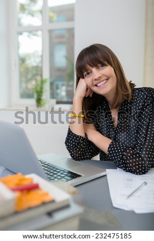 Happy vivacious businesswoman sitting at her desk in the office giving the camera a lovely warm smile