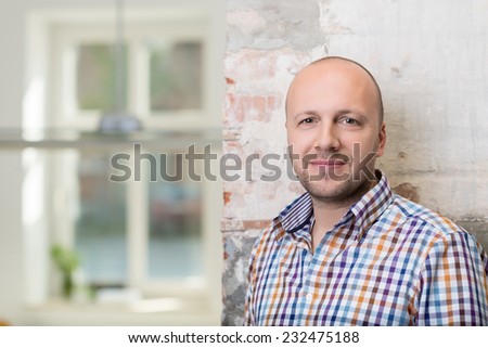 Balding middle-aged man in a checked shirt standing against a painted brick wall looking at the camera with a friendly smile, with copyspace