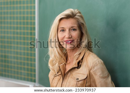 Attractive stylish middle-aged woman teacher standing in front of the class blackboard looking thoughtfully at the camera with a smile