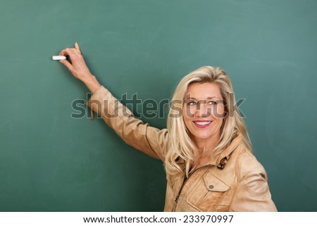 Attractive stylish blond woman teacher writing on a blank blackboard in class turning to watch her students