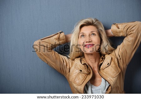 Attractive mature woman standing thinking with her hands behind her head leaning on a dark background looking into the air with a smile