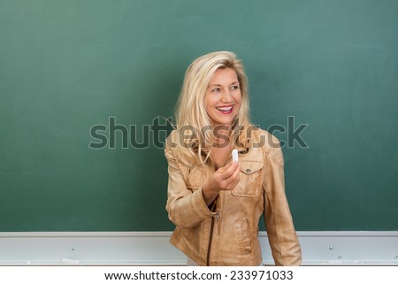 Vivacious teacher offering the chalk holding it up in her hand as she stands in front of a blank blackboard inviting a student to the front of the class