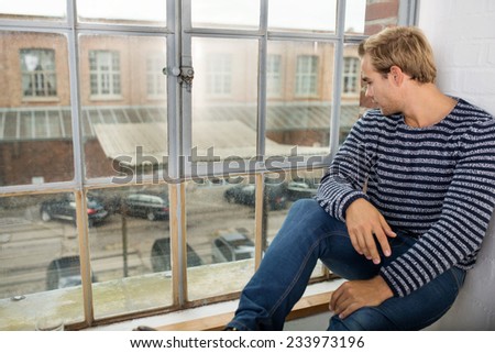 Young man sitting on the windowsill in his apartment looking out of a window at the parking lot and urban street below