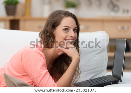 Pretty woman using a laptop on a sofa smiling as she lies looking up into the air with a dreamy expression