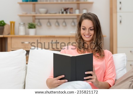 Young woman spending a relaxing day at home sitting on a sofa with a good book smiling with pleasure and enjoyment