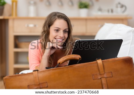 Smiling young woman on vacation leaning on an old leather suitcase reading a tablet computer as she keeps in touch with friends and family