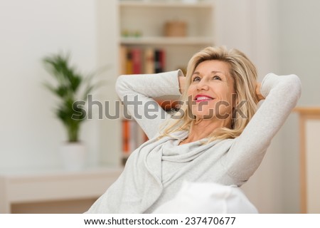 Woman relaxing at home with a contented smile as she leans back in her chair with her hands behind her head looking up into the air with a look of pleasure