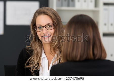 Close up Pretty Young Business Executive with Long Blond Hair and Eyeglasses Smiling at the Camera while Talking to her Female Co-worker at the Office.