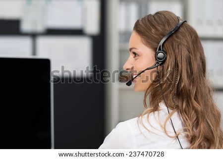 Young businesswoman in a call center or client services desk wearing a headset and smiling as she assists a customer
