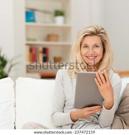 Smiling attractive middle-aged blond woman sitting on a couch at home holding a tablet-pc and looking at the camera