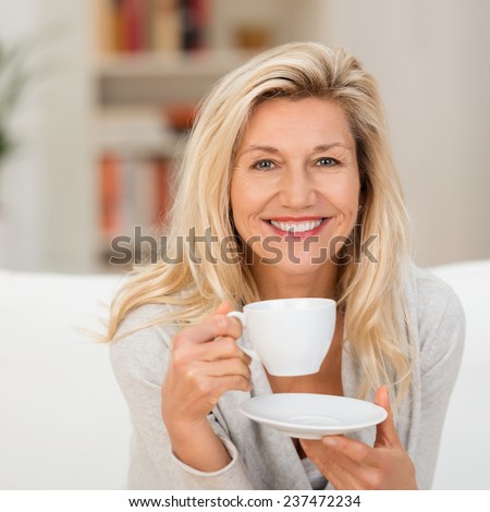 Happy vivacious middle-aged blond woman holding a cup of tea or coffee looking at the camera with a beaming warm smile