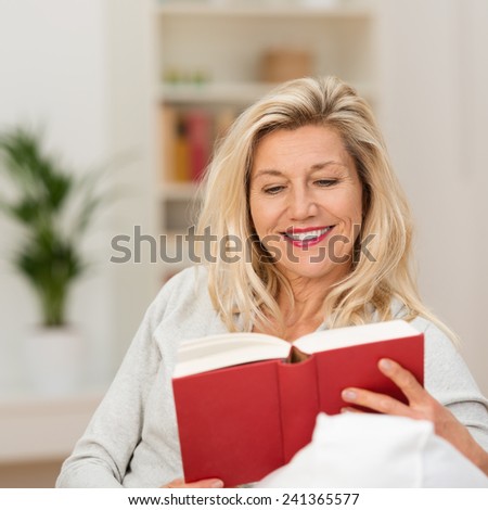 Attractive blond middle-aged woman enjoying reading a book sitting on the sofa in her living room smiling as she reads