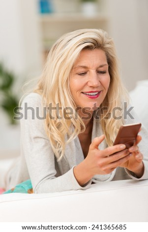 Middle-aged woman reading a text message on her mobile phone with a look of smiling anticipation as she lies on a sofa at home