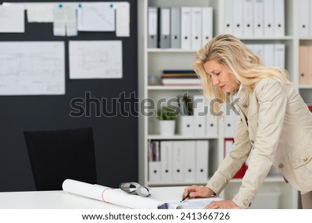 Businesswoman working in her office standing at her desk reading a document with a rolled up poster or architectural drawing alongside her, shelving background with office binders