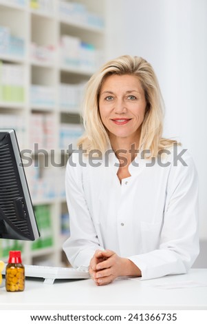 Attractive middle-aged woman pharmacist standing at the counter in the pharmacy behind a computer looking at the camera