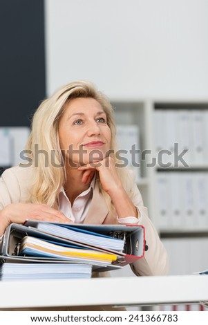 Attractive middle-aged businesswoman sitting daydreaming at her desk resting her chin on her hand staring upwards with a smile