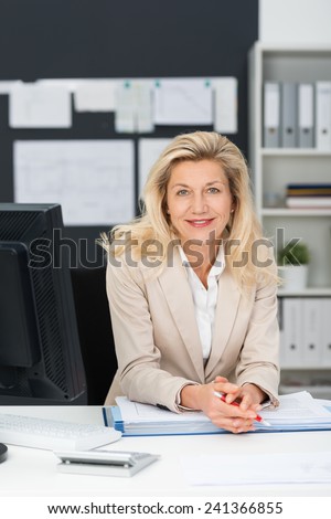 Friendly stylish middle-aged businesswoman leaning on a folder on her desk in the office smiling at the camera