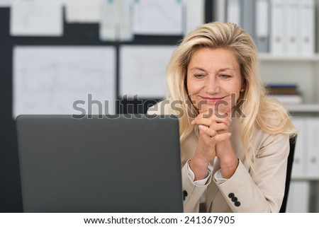 Pleased attractive middle-aged businesswoman sitting at her desk with a broad smile reading information on her laptop screen