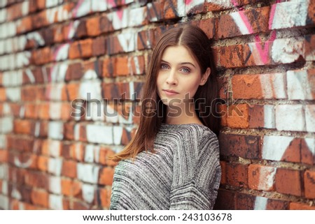 Pretty young city girl standing against an exterior brick wall sprayed with graffiti looking at the camera with a serious expression