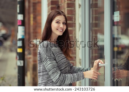 Smiling attractive young female student entering a commercial building looking at the camera as she pushes open the glass door