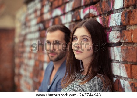 Thoughtful young couple standing leaning against a face brick wall covered in graffiti watching to the left of the frame
