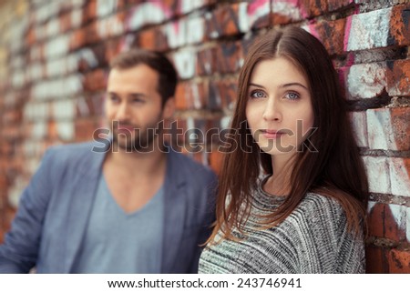 Attractive young woman with a serious expression standing against a graffiti covered brick wall in an urban street alongside her husband or boyfriend looking at the camera