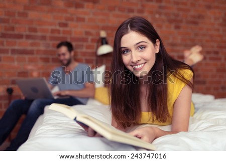 Woman relaxing on the weekend with a good book lying on her stomach on the bed looking at the camera with a smile as her husband works on a laptop in the background
