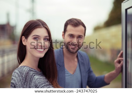 Young man and woman on a train station standing checking the time table in a notice board and looking at the camera with friendly smiles, close up of their faces