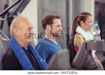 Group of diverse people in a gym working out together on the equipment with an elderly man and young woman with focus to the smiling face of a young man in the centre, in a health and fitness concept