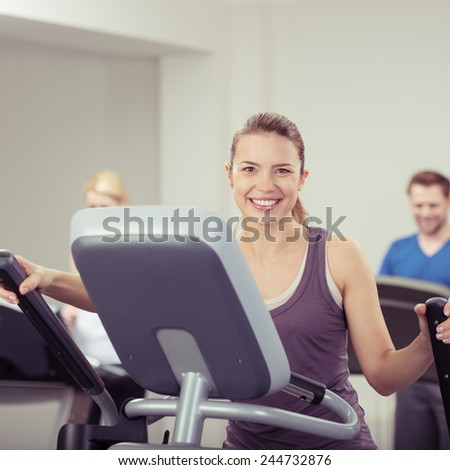 Fit vivacious young woman at the gym working out on the equipment while looking at the camera with a warm friendly smile
