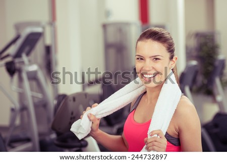 Happy vivacious young woman in a gym standing surrounded by equipment with a towel around her shoulders smiling at the camera, head and shoulders in a fitness concept