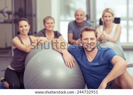 Smiling handsome young man in pilates class at the gym sitting leaning on his gym ball with the rest of the diverse group gathered behind him