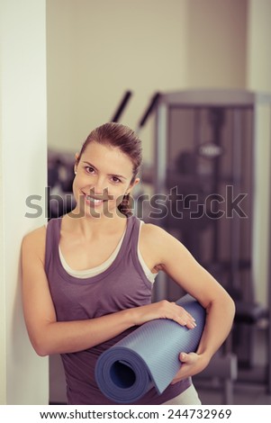 Attractive young woman carrying an exercise mat rolled up under her arm in a gym standing leaning against a wall looking at the camera with a friendly smile in a healthy lifestyle concept