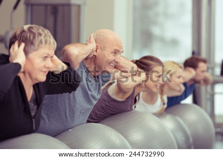 Happy elderly couple exercising in a pilates class at the gym with three other younger people toning and strengthening their muscles using gym balls, focus to the senior man and woman