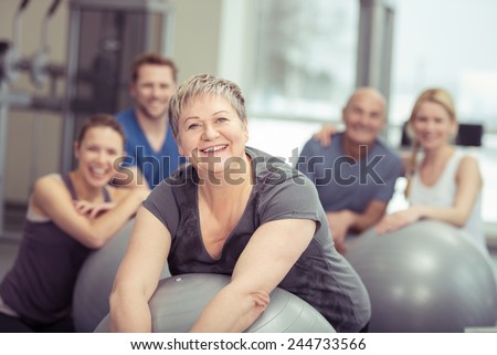 Smiling senior woman enjoying pilates class at the gym posing leaning on her ball smiling at the camera with the class behind