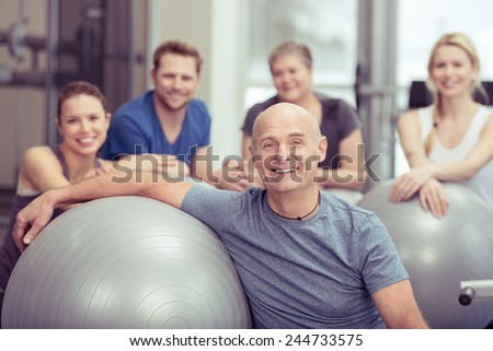 Smiling happy fit senior man in a gym class with a group of diverse people leaning on a pilates ball looking at the camera in a healthy lifestyle concept
