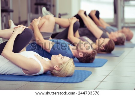 Diverse group of young and older people exercising at the gym doing leg flexes as they lie on their backs on their mats in a receding row, focus to an attractive young blond girl in the foreground