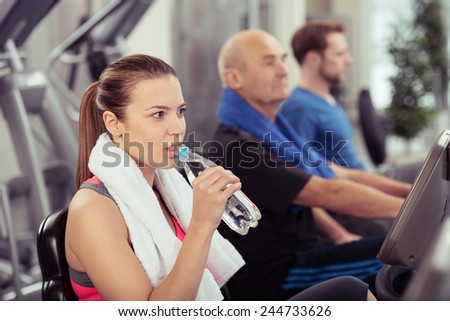 Young woman pausing during her workout to drink fresh bottled water in a gym with other people on equipment in the background