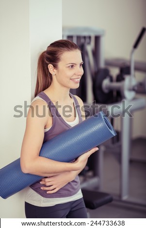Fit athletic young woman carrying an exercise mat rolled up under her arm at the gym standing sideways looking to the right of the frame with a smile