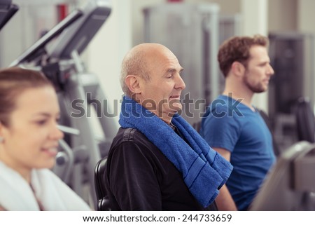 Senior man working out in a gym flanked by a young man and woman with focus to the older man in a health and fitness concept