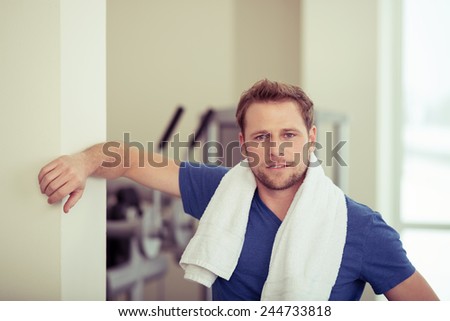 Handsome fit young man in a gym posing leaning against a wall with a towel around his neck looking at the camera